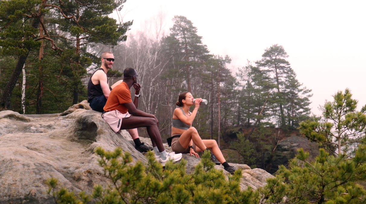 Group of active young adults resting on a scenic mountain rock ledge, surrounded by pine trees and forest landscape, with one woman drinking water after a hike.