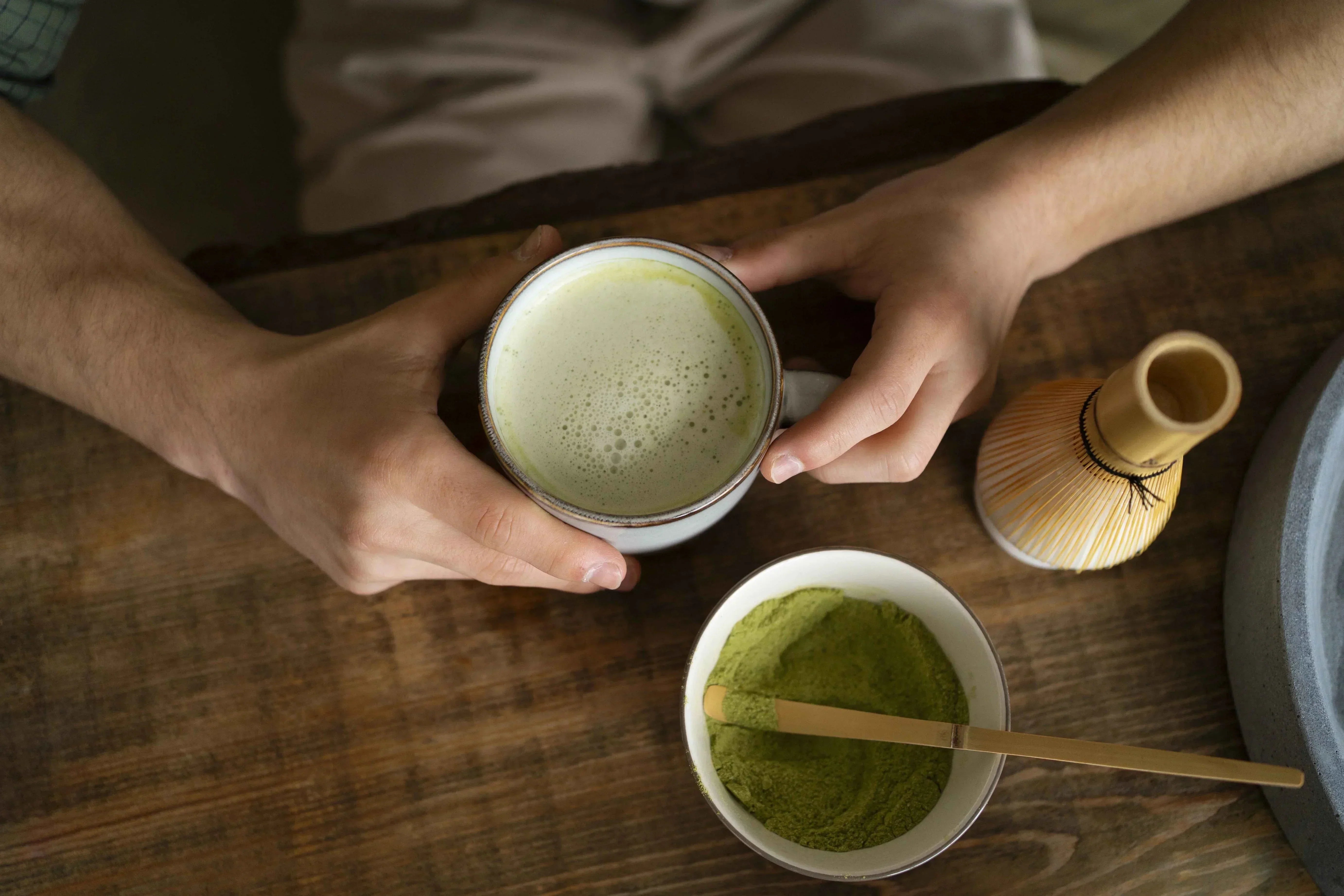 Person holding a cup of frothy matcha with matcha powder and whisk nearby.