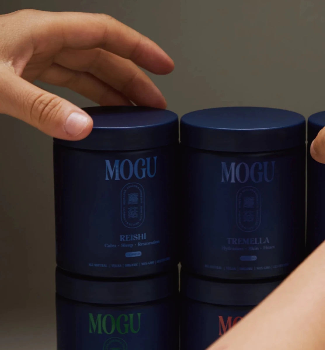 Reishi capsules bottle among blue containers labeled 'MOGU' with a hand reaching towards them on a reflective surface.