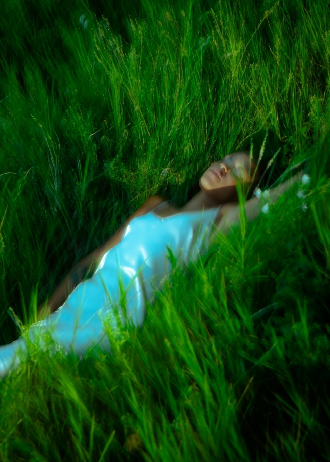 Alt text: Woman in a white satin dress lying peacefully in tall green grass, surrounded by wild plants, evoking a serene, dreamlike atmosphere.