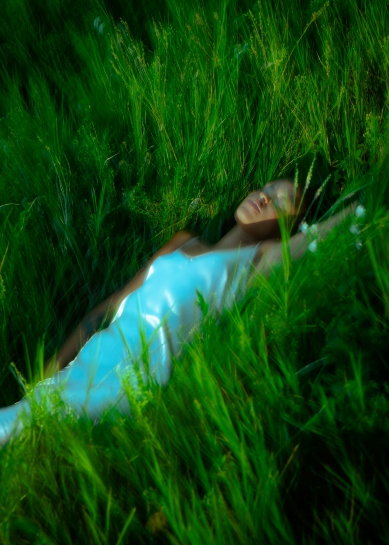 Alt text: Woman in a white satin dress lying peacefully in tall green grass, surrounded by wild plants, evoking a serene, dreamlike atmosphere.