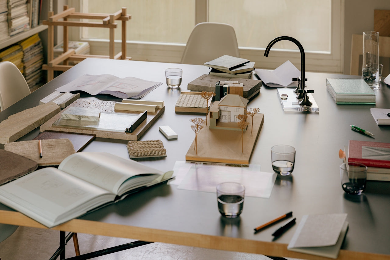 Architect's workspace with scale model house, material samples, design blueprints, and sketchbooks on large modern table in well-lit studio.